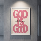 God Is Good pink typography poster with black “all the time” script on cream, shown in a white frame against a gray wall.
