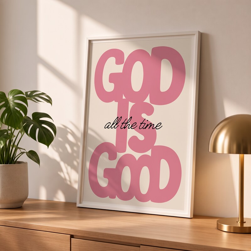 God Is Good pink Christian poster in a styled tabletop mockup beside a plant and brass-colored lamp.