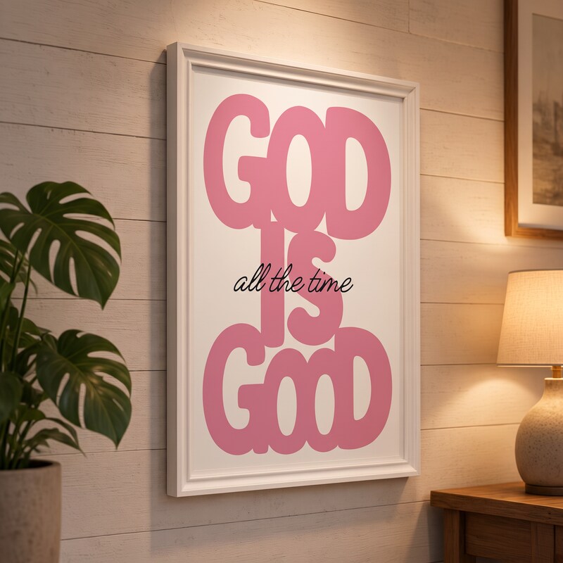 God Is Good pink Christian poster displayed in a white frame on a paneled wall beside a plant and lamp.