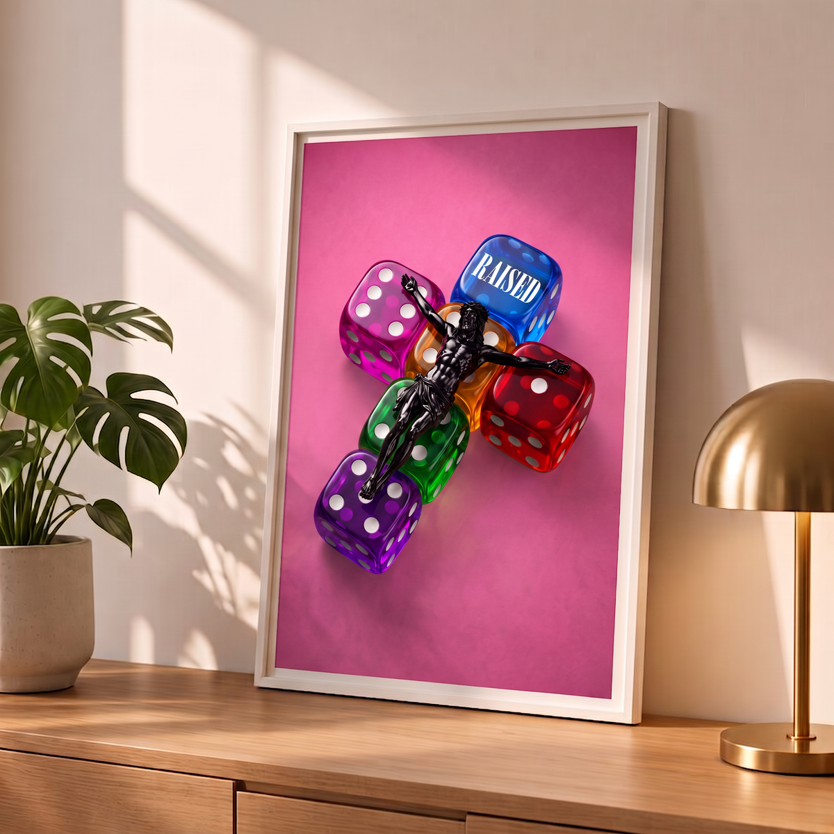 RAISED pink Jesus poster in a styled tabletop mockup beside a plant and brass-colored lamp.