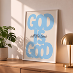 God Is Good blue Christian poster in a styled tabletop mockup beside a plant and brass-colored lamp.