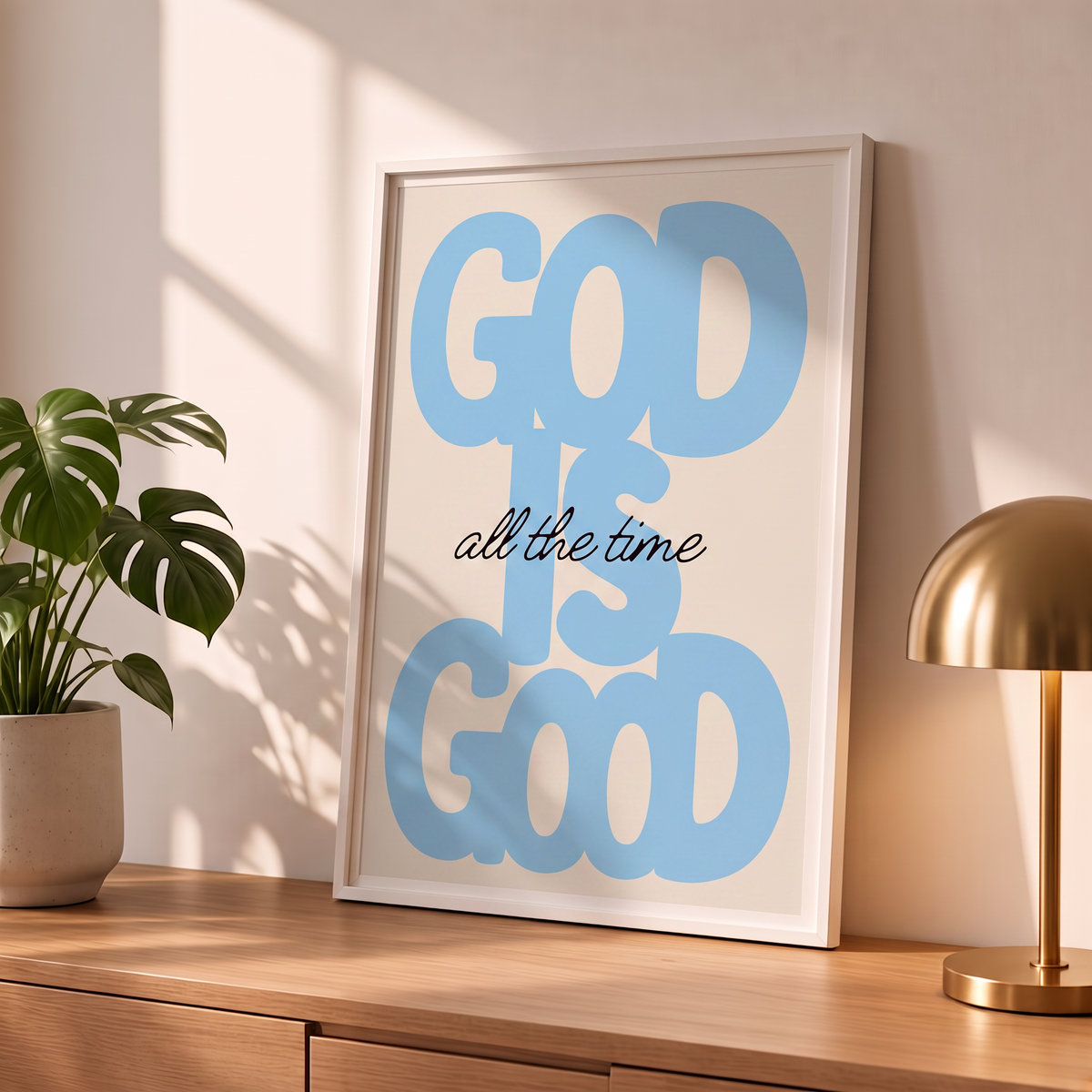 God Is Good blue Christian poster in a styled tabletop mockup beside a plant and brass-colored lamp.
