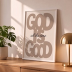 God Is Good brown Christian poster in a styled tabletop mockup beside a plant and brass-colored lamp.