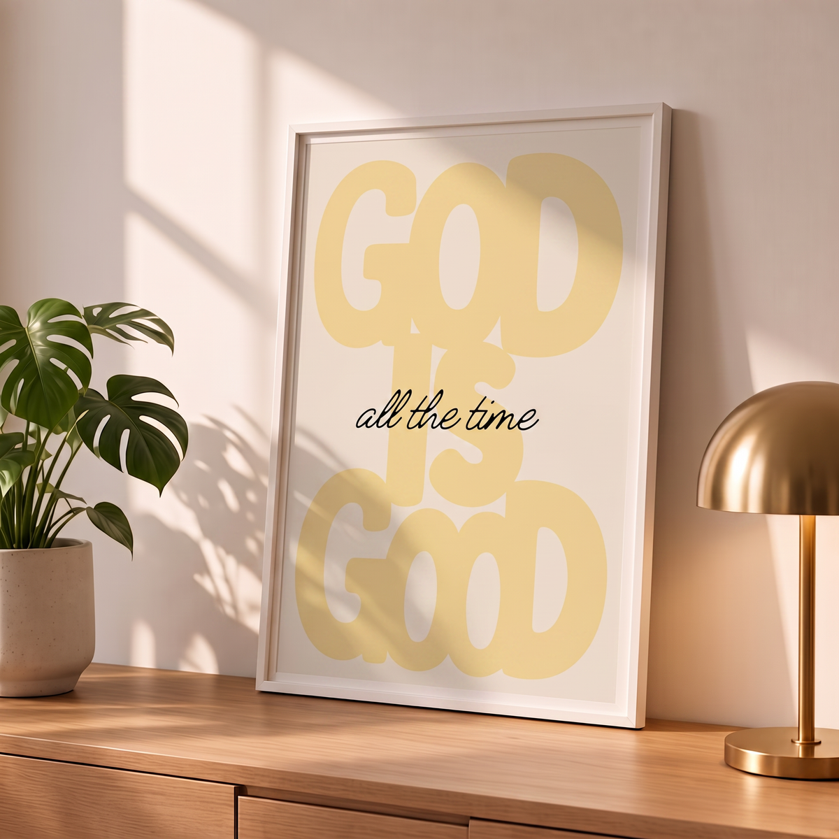 God Is Good yellow poster in a styled tabletop mockup beside a plant and brass-colored lamp.