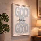 God Is Good grey Christian poster displayed in a white frame on a paneled wall beside a plant and lamp.